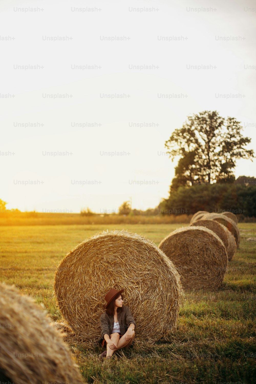 Stylish girl relaxing at hay bale in summer field in sunset. Young woman in hat sitting at haystack, atmospheric tranquil moment. Countryside slow life