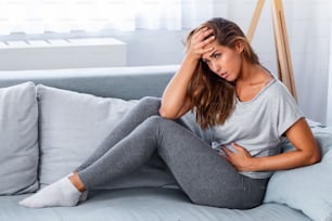 View of young woman suffering from stomachache on sofa at home. Woman sitting on bed and having stomach ache. Young woman suffering from abdominal pain while sitting on sofa at home