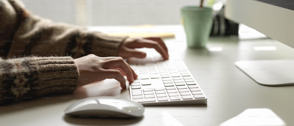 Vista lateral de la empleada que escribe en el teclado de la computadora en el escritorio mínimo de la oficina con el libro de horarios y la taza