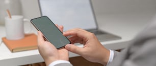 Close up view of male entrepreneur hands using  mock-up smartphone while sitting at white office desk with office supplies