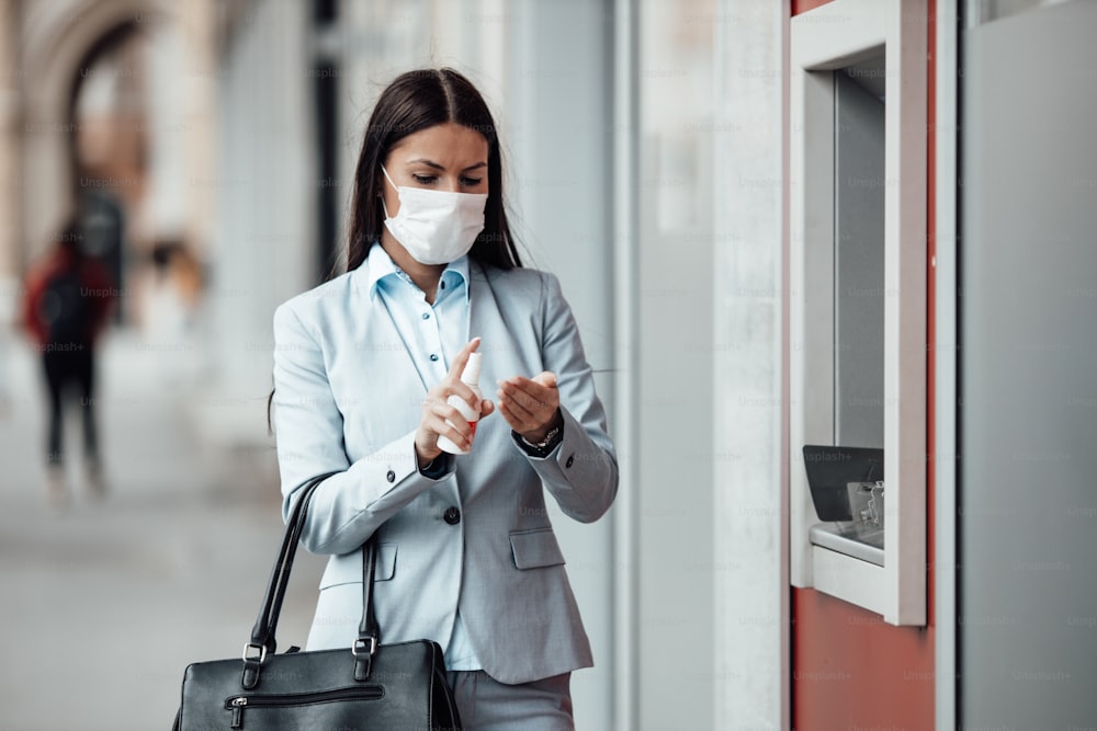 보호 마스크를 쓴 우아한 비즈니스 여성이 도시 거리에 서서 ATM 기계를 사용한 후 알코올 스프레이를 사용하여 손을 소독합니다. 코로나 또는 Covid-19 바이러스 전염병 예방 및 의료 개념.
