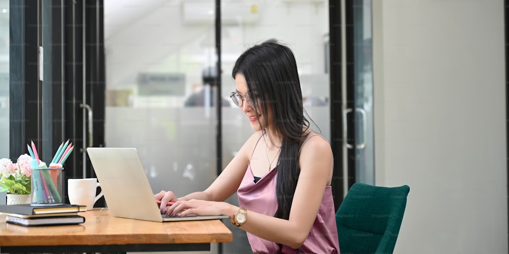 A beautiful woman is typing on a computer laptop while sitting at the wooden working desk.