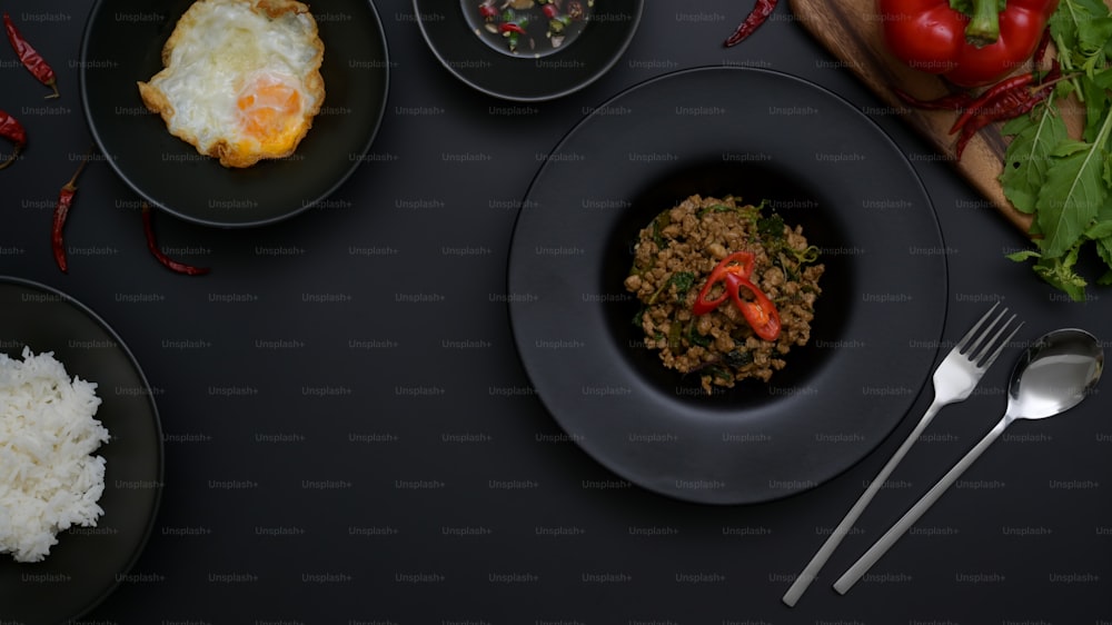 タイの伝統料理、豚ひき肉のバジル炒め(パッカプラオ)、黒い陶器の皿に盛られた目玉焼き、黒いテーブルに銀食器とコピースペース