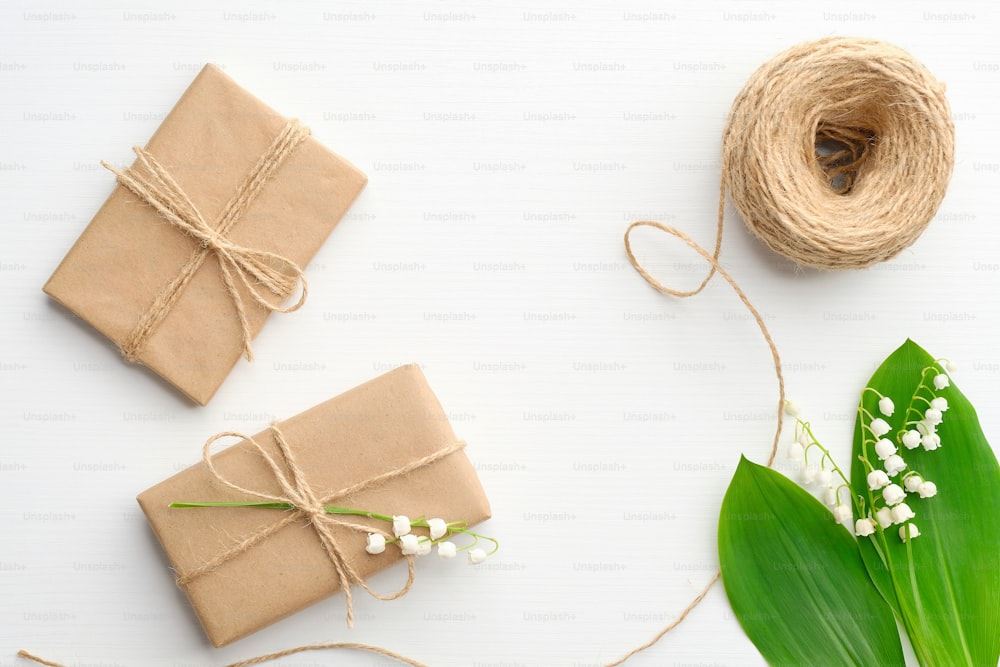 Geschenk- oder Geschenkboxen eingewickelt in Kraftpapier, Bindseil und Blumenmaiglöckchen auf weißem Tisch von oben. Hochzeits- oder Jubiläumskonzept