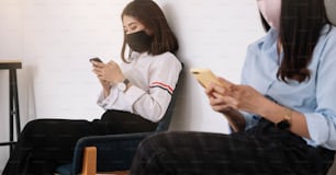 Dos jóvenes asiáticas que chatean en un teléfono inteligente y usan mascarilla a una distancia sentada protegen de los virus COVID-19 para el distanciamiento social por riesgo de infección