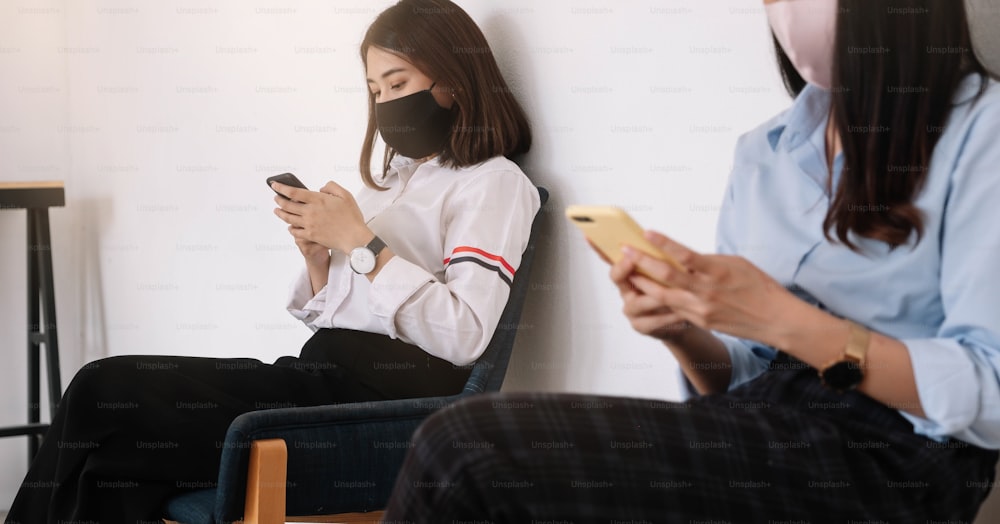 Two Asian young woman chatting in smartphone and wearing mask sitting distance protect from COVID-19 viruses for social distancing for infection risk