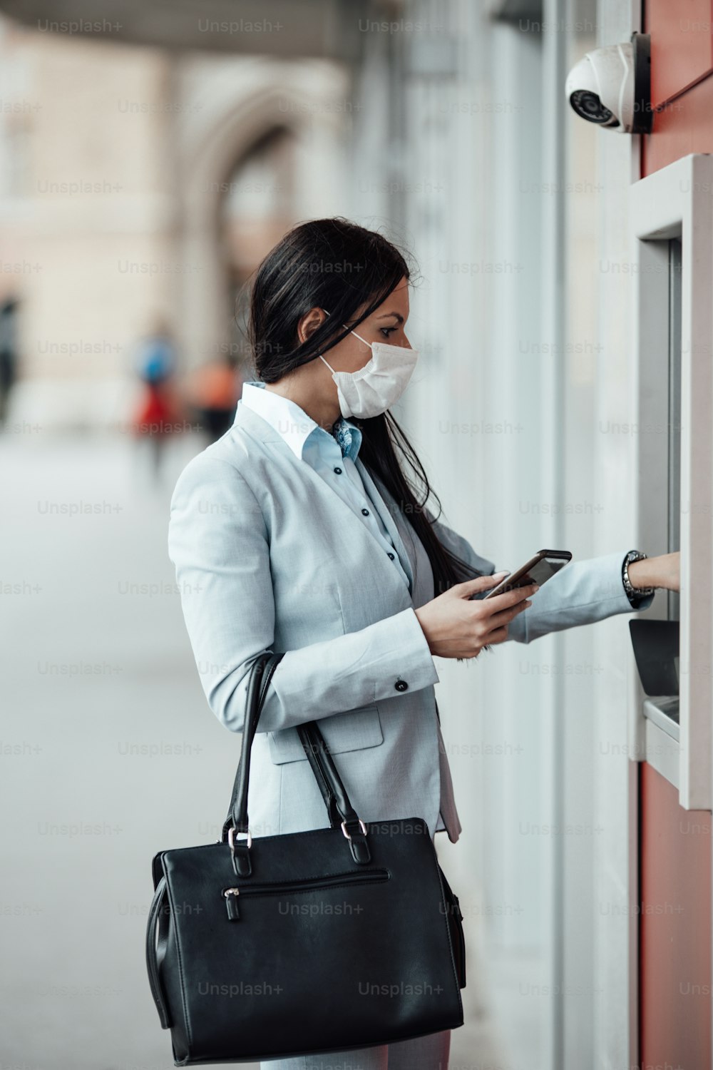 防護マスクを着用したエレガントなビジネスウーマンが街の通りに立ち、ATM機を使って現金を引き出します。コロナウイルスまたはCovid-19ウイルスのパンデミックのコンセプト。