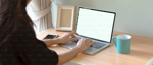 Cropped shot of female working at home with mock-up laptop, smartphone, mug and mock-up frame on wooden worktable in bed room