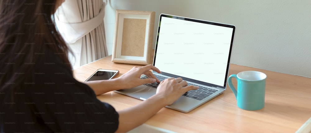 Cropped shot of female working at home with mock-up laptop, smartphone, mug and mock-up frame on wooden worktable in bed room