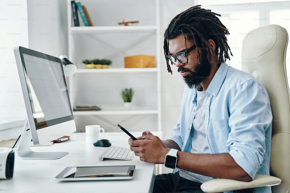 Hübscher junger afrikanischer Mann in Brillen mit Smartphone während der Arbeit im Büro