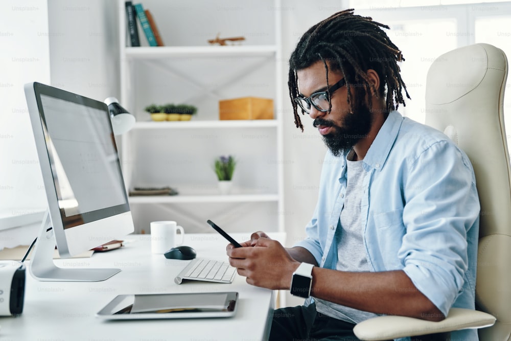 Joven africano guapo con gafas usando un teléfono inteligente mientras trabaja en la oficina