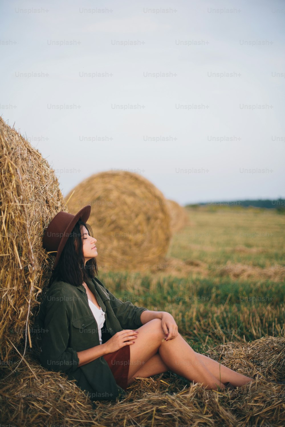 Stylish girl relaxing at hay bale in summer field in sunset. Young woman in hat sitting at haystack, atmospheric tranquil moment. Countryside slow life