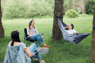 Soziale Distanzierung. Kleine Gruppe von Menschen, die Gespräche beim Picknick mit sozialer Distanz im Sommerpark genießen. Freunde chillen in Hängematte und Stühlen zwischen Bäumen. Neue Normalität, Sicherheitsversammlungen