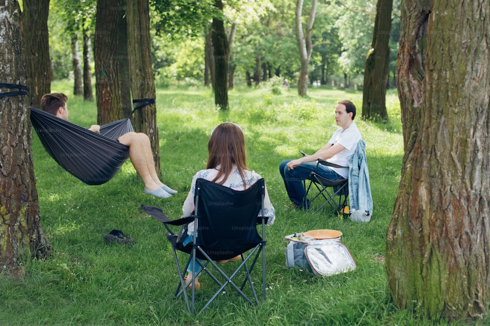 Small group of people enjoying conversation at picnic in accordance with social distancing in summer park. Friends chilling in hammock and on chairs among trees
