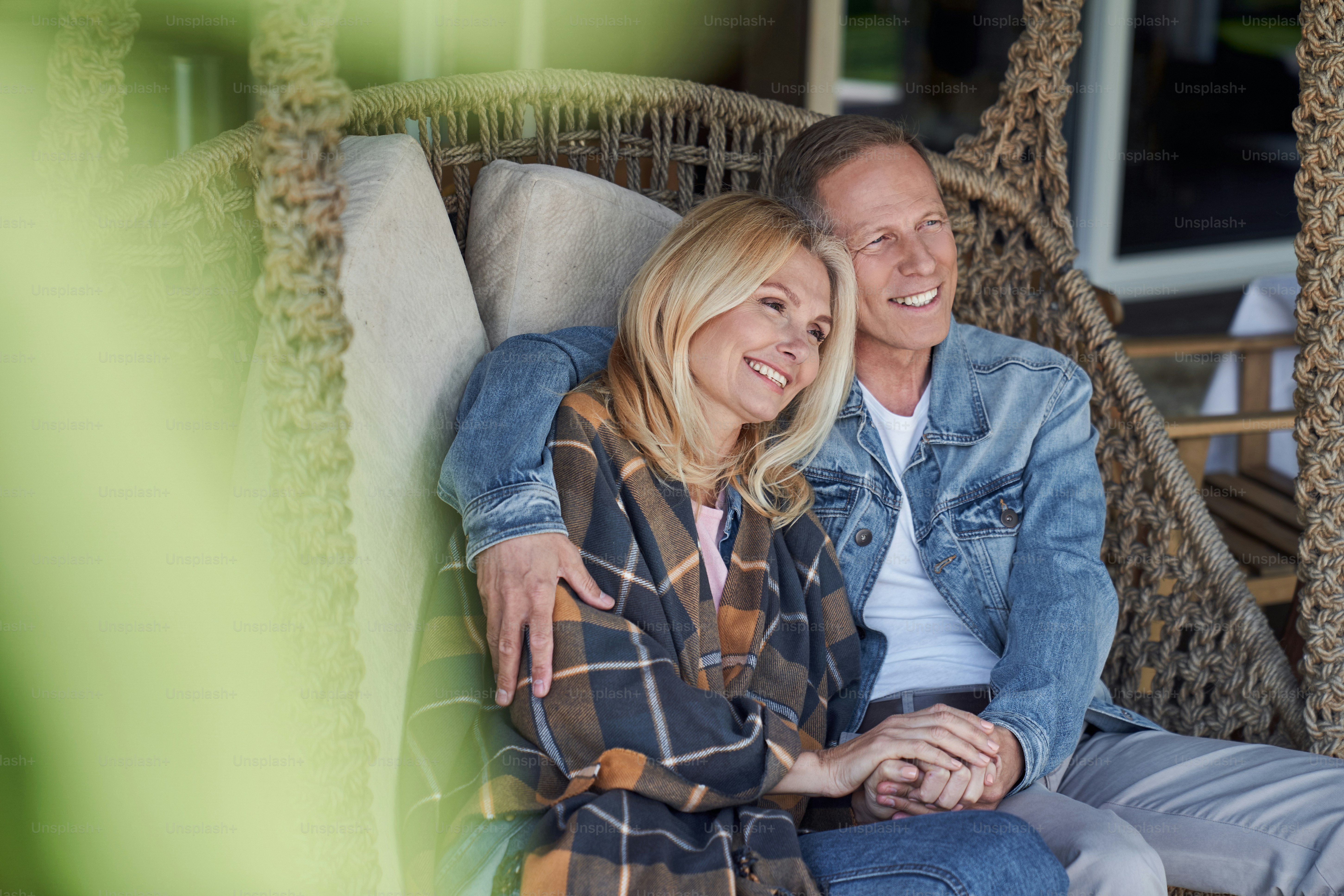 Lächelndes Liebespaar sitzt in Korbschaukeln auf dem Land und hält sich an den Händen