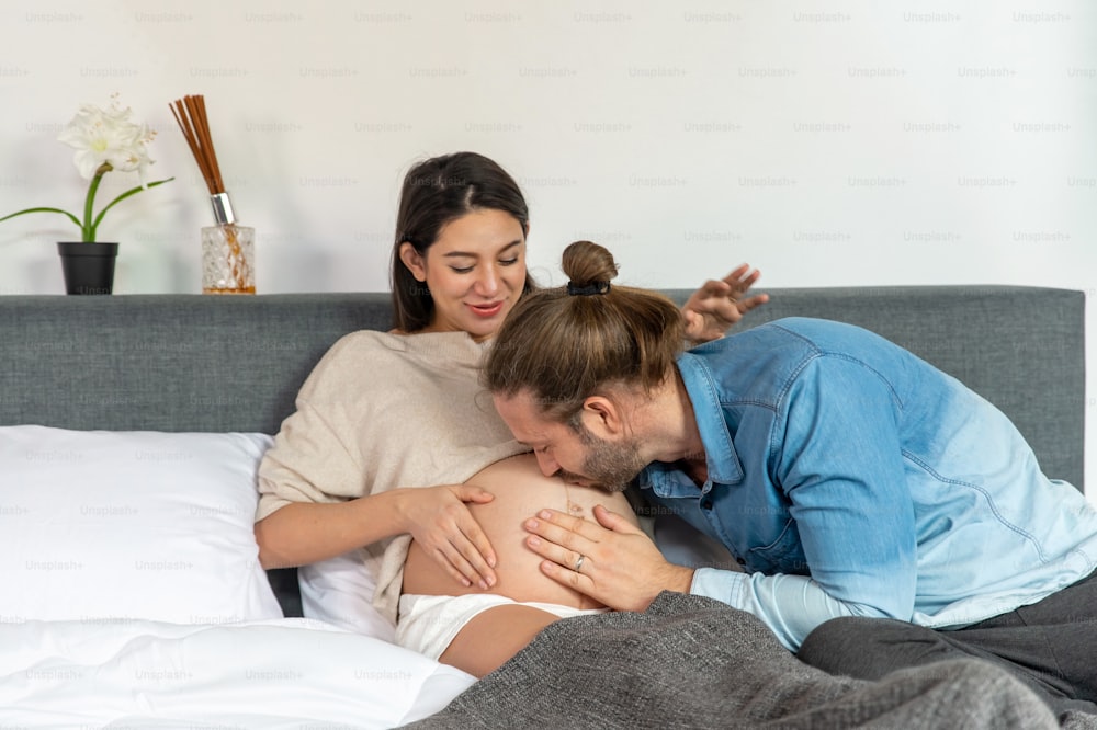 Caucasian man touching and kissing his pregnant wife's tummy on the bed.
