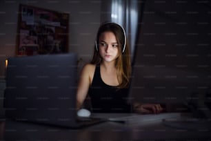 Frontansicht eines gelangweilten jungen Mädchens mit Kopfhörern und Computer, das drinnen sitzt, Online-Chat-Konzept.