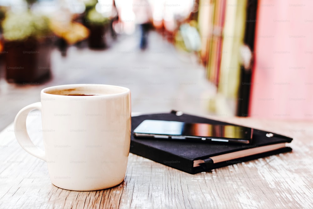 taza de café y teléfono inteligente al aire libre en una mesa en una cafetería o cafetería con fondo borroso