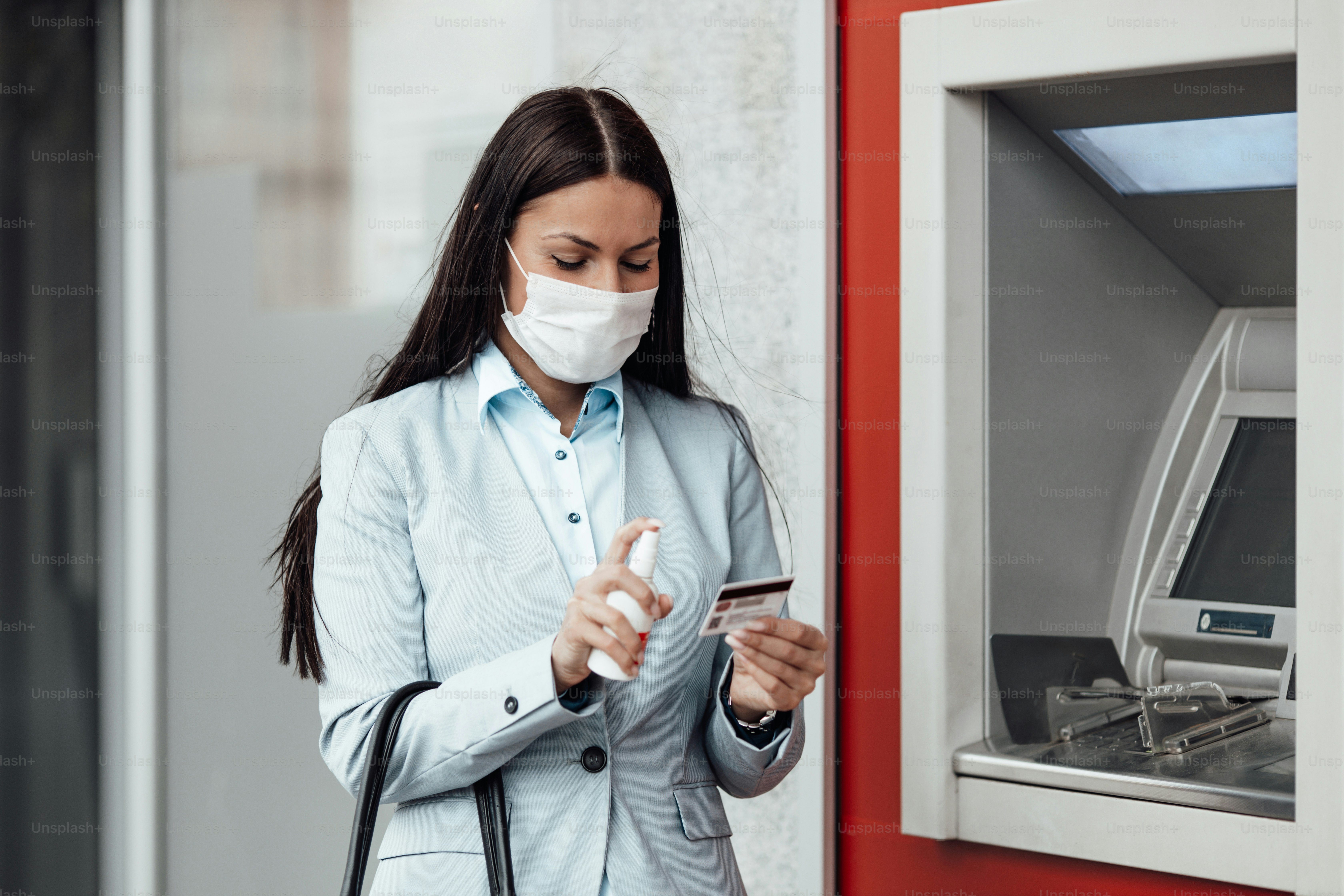 Elegant business woman with protective mask standing on city street and using alcohol spray to disinfect her hands after use of ATM machine. Corona or Covid-19 virus pandemic prevention and healthcare concept.