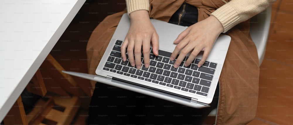 Vue aérienne de mains féminines tapant sur un ordinateur portable sur ses genoux alors qu’elle est assise sur une chaise de bureau dans une salle de bureau