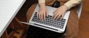 Overhead-Aufnahme von weiblichen Händen, die auf ihrem Schoß auf dem Laptop tippen, während sie auf einem Bürostuhl im Büroraum sitzen