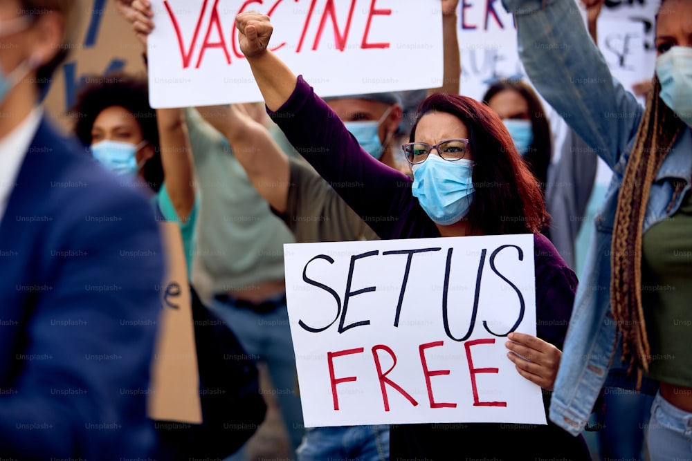 Female activist wearing protective face mask while protesting with group of people and supporting anti-vaccination movement.
