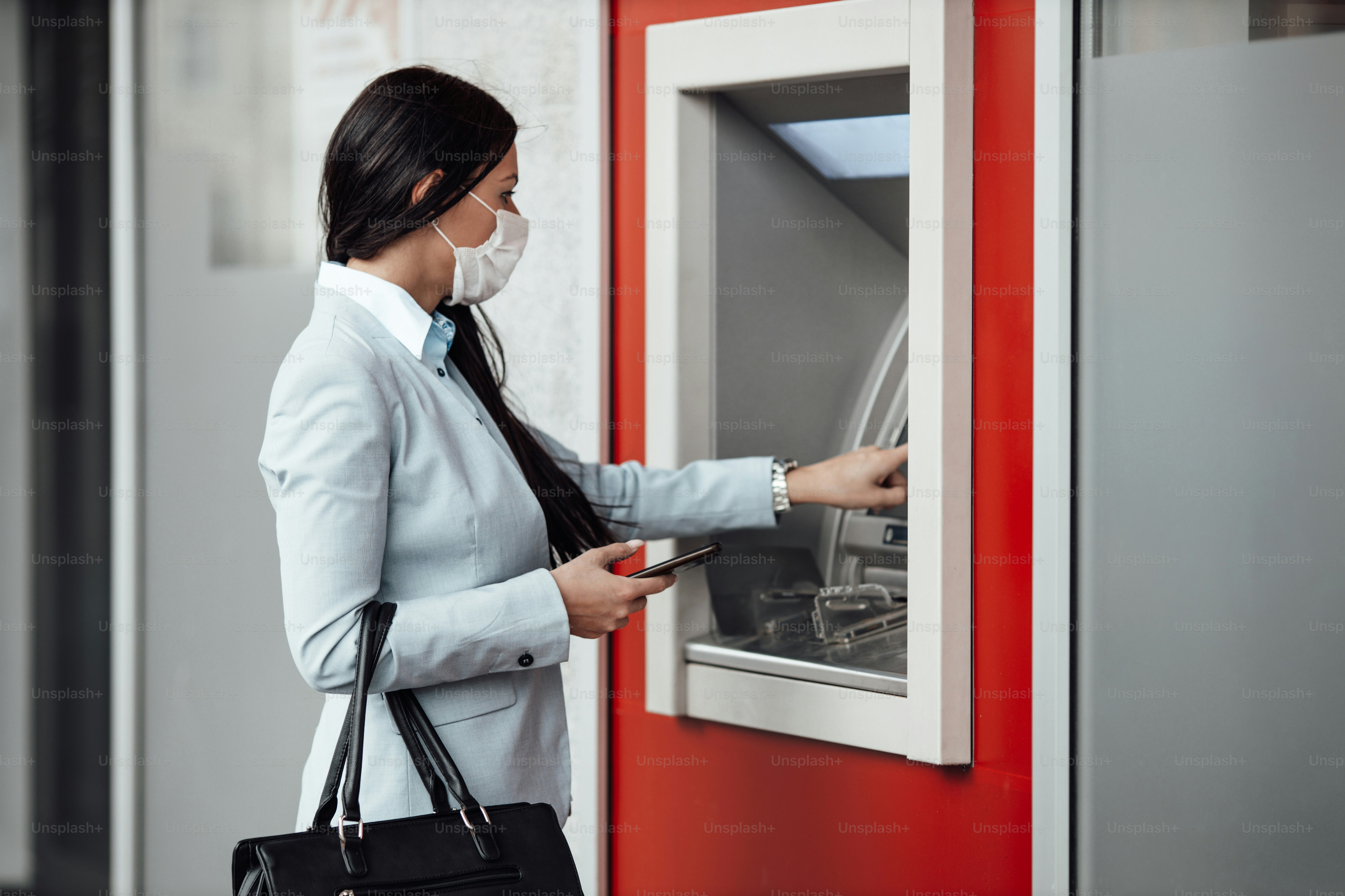 Elegant business woman with protective mask standing on city street and using ATM machine to withdraw cash. Corona or Covid-19 virus pandemic concept.