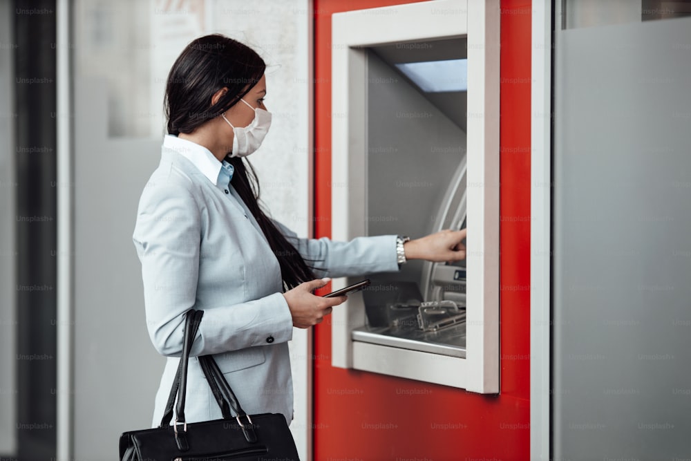 Elegante Geschäftsfrau mit Schutzmaske, die auf der Stadtstraße steht und am Geldautomaten Bargeld abhebt. Corona- oder Covid-19-Virus-Pandemie-Konzept.