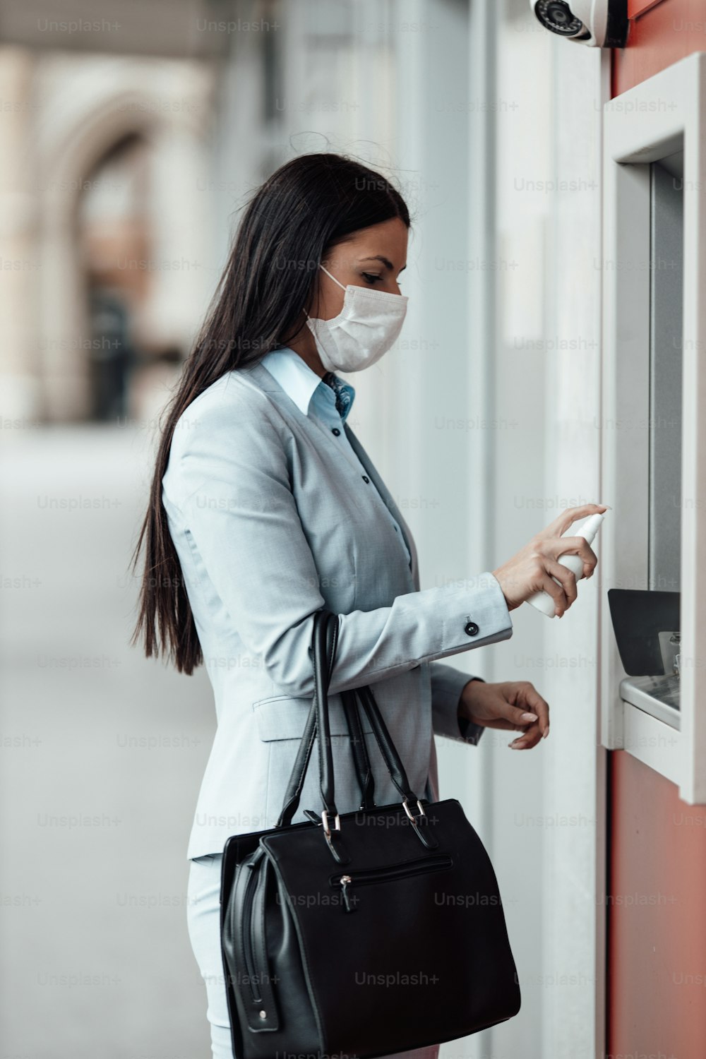 보호 마스크를 쓴 우아한 비즈니스 여성이 도시 거리에 서서 ATM 기계를 사용한 후 알코올 스프레이를 사용하여 손을 소독합니다. 코로나 또는 Covid-19 바이러스 전염병 예방 및 의료 개념.