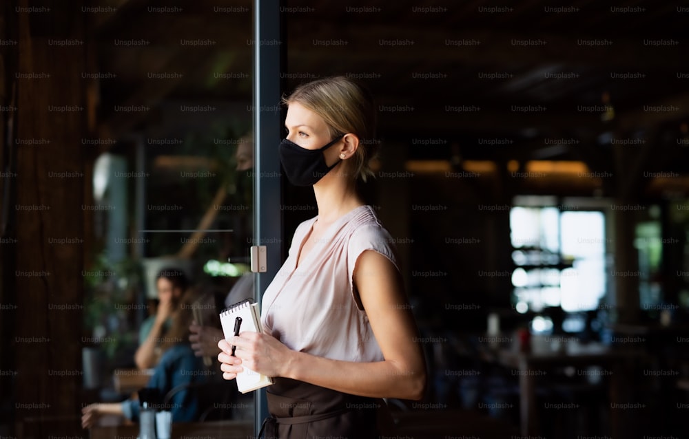 Side view of waitress with face mask standing at the door in restaurant, holding order pad.