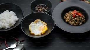 Thailändisches traditionelles Essen, gebratenes Schweinehackfleisch mit Basilikum (Pad ka prao), Reis, Spiegelei und Chili-Fischsauce auf schwarzen Keramiktellern im thailändischen Restaurant