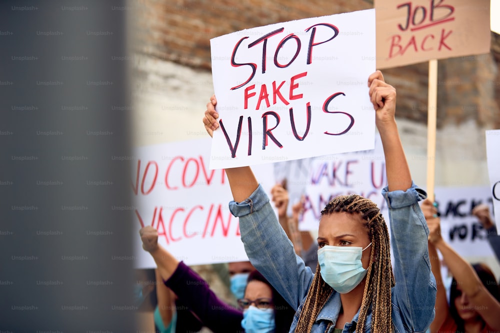 Una mujer caucásica con una máscara protectora mientras sostiene un cartel de "Detengan el virus falso" durante una protesta en las calles de la ciudad.