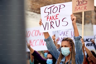 Caucasian woman wearing protective face mask while holding 'Stop fake virus' sign during a protest on city streets.