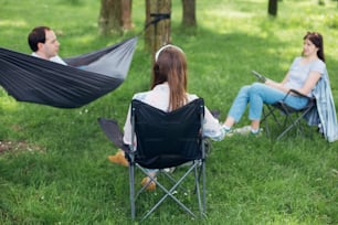 Social distancing. Small group of people enjoying conversation at picnic with social distance in summer park. Friends chilling in hammock and chairs among trees. New normal, safety gatherings