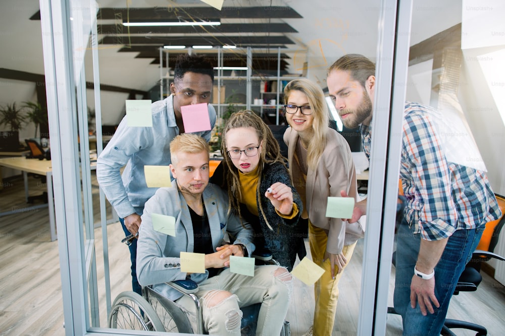 Inclusion for disabled people. Group of young multiracial business people with one handicapped man in wheelchair, discussing ideas in front of glass wall using post it notes and stickers.