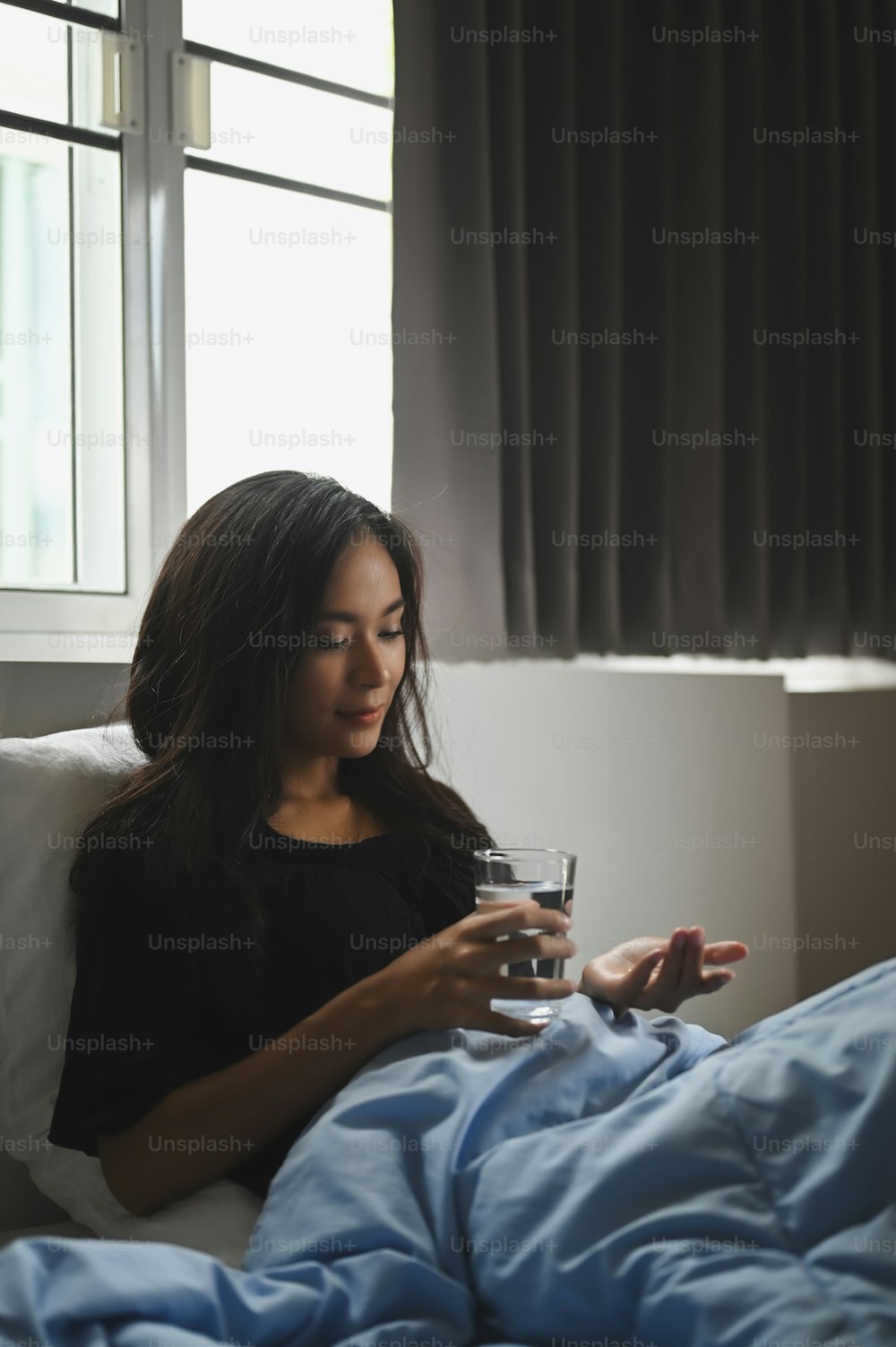 An ill woman is holding a glass of water and consuming a pill at the bed.