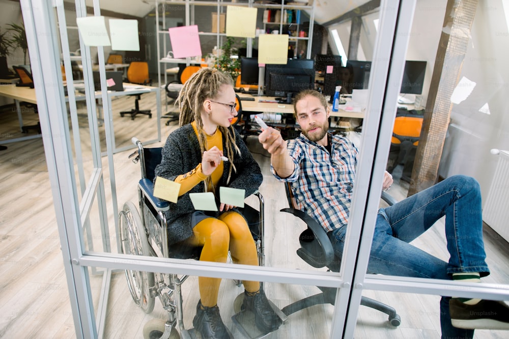 Two business colleagues, pretty disabled blond girl with long dreadlocks and handsome bearded man, brainstorming together near a glass wall with sticky notes in a modern office