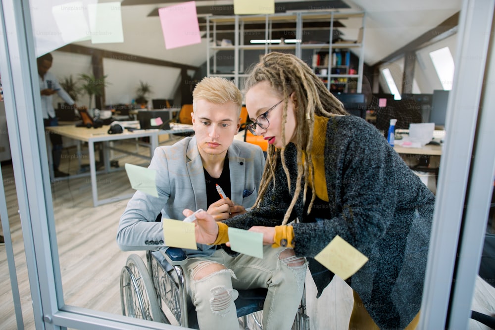 Two happy creative concentrated businesspeople, disabled man in wheelchair and pretty blond girl with dreadlocks, brainstorming in office, placing sticky notes with ideas on glass wall.