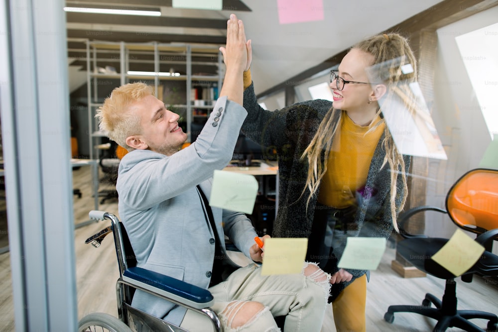 Business concept, successful disabled people at work. Happy successful young man in wheelchair giving a high five to his pretty female coworker with dreadlocks, laughing and cheering their success.