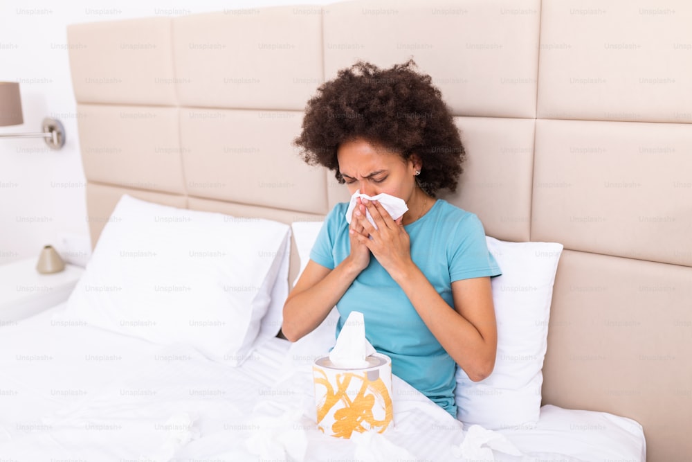 Sick black millennial girl sit on bed feel unhealthy blowing running nose, ill african American young woman suffering from rhinitis snuffles, having respiratory infection, get flue need medication