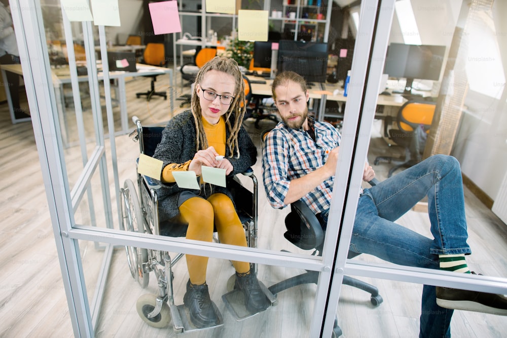Smiling young disabled hipster businesswoman with blond dreadlocks, sitting in wheelchair, reading sticky notes on a glass wall and writing her ideas while brainstorming with male coworker in office.