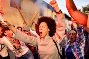 Angry crowd of people taking a part in public demonstrations. Focus is on African American woman shouting with raised fist.