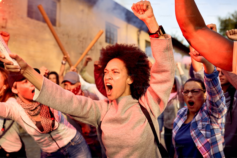 Angry crowd of people taking a part in public demonstrations. Focus is on African American woman shouting with raised fist.