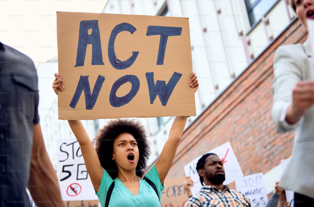 Low angle view of young black woman holding act now sign and shouting while taking a part in demonstrations on city streets.