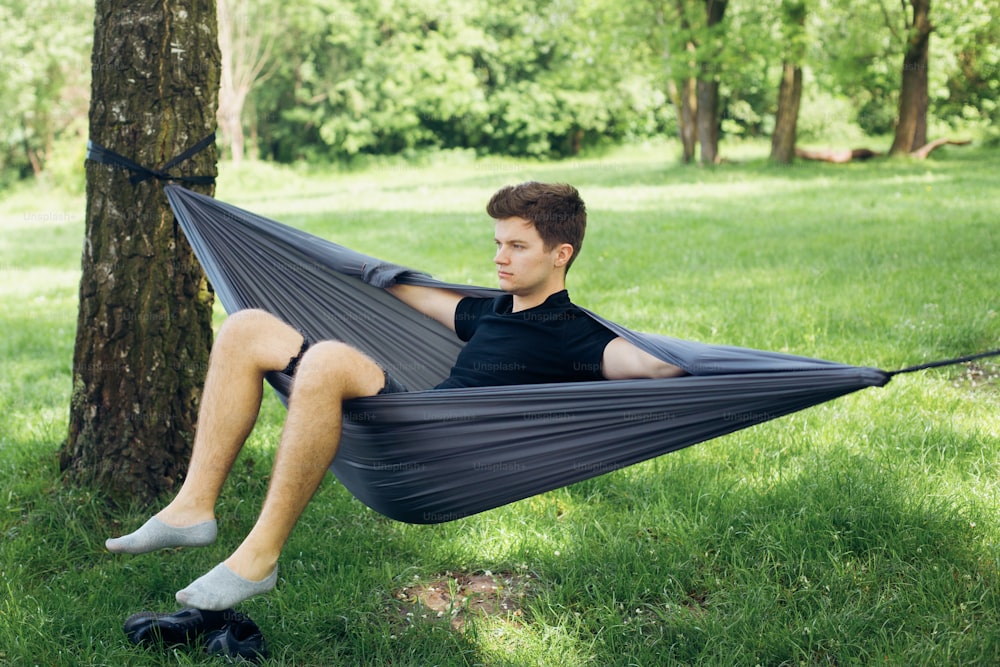 Young man chilling in hammock among trees. Social distancing. Small group of people enjoying conversation at picnic with social distance in summer park.