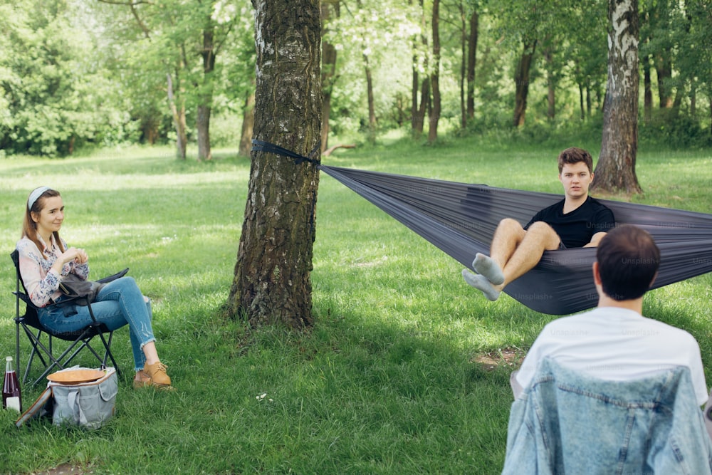 ソーシャルディスタンシング。夏の公園でソーシャルディスタンスを保ったピクニックで会話を楽しむ少人数のグループ。ハンモックや木々の間の椅子でくつろぐ友人たち。新世界での安全集会