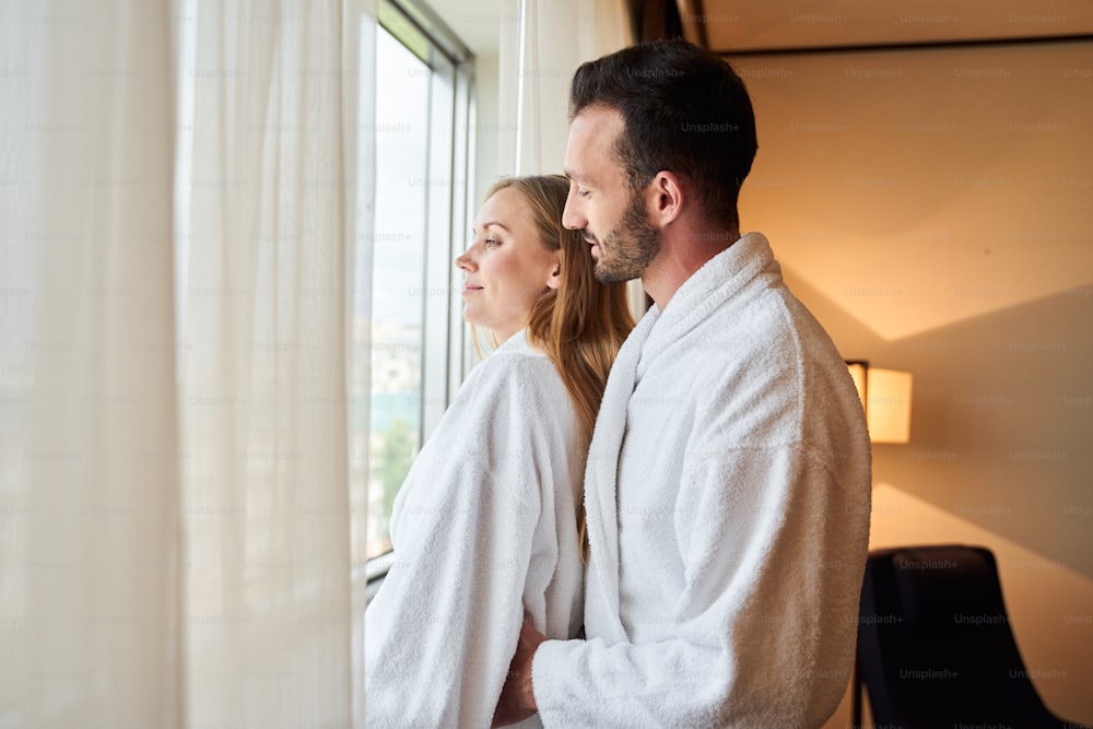 Caring bearded man is hugging young beloved wife while standing at window and enjoying view