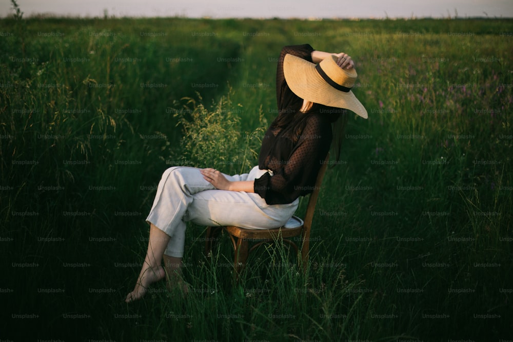 Vida lenta. Elegante menina elegante no chapéu de palha sentado na cadeira rústica no prado verde do verão na luz do sol da noite. Mulher jovem elegante relaxando no campo, momento tranquilo. Imagem criativa