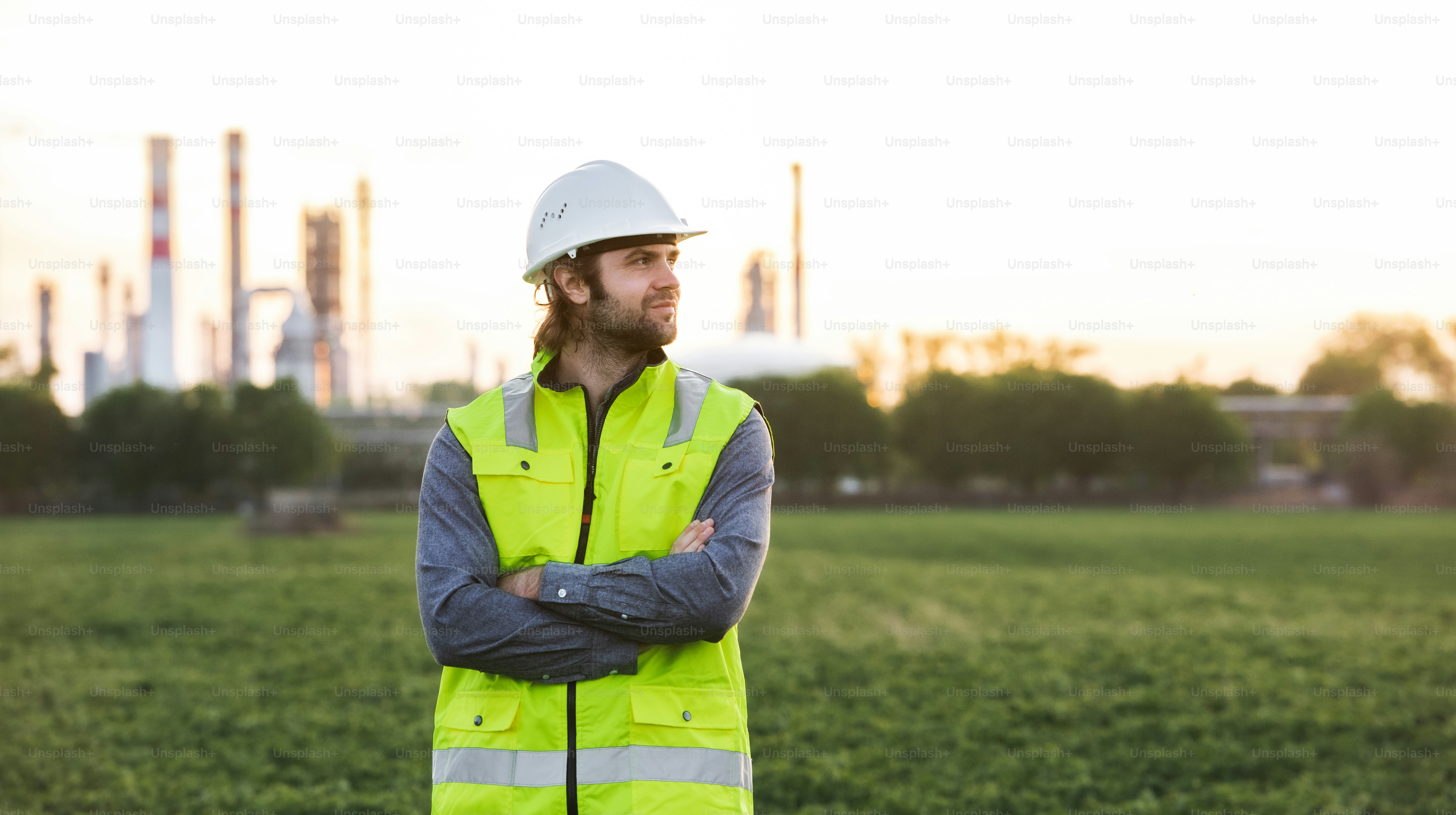 Portrait of young engineer standing outdoors by oil refinery, arms crossed.