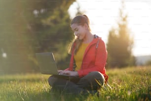 Vista lateral de una mujer joven feliz que usa una computadora portátil en la naturaleza del verano, concepto de oficina al aire libre.